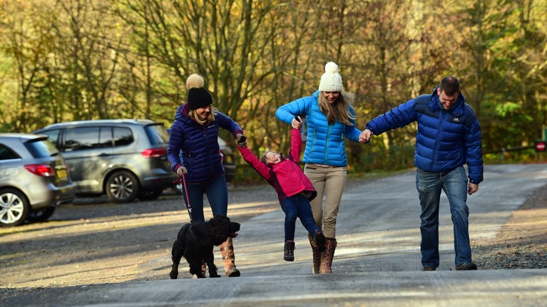 Visitors walking away from a car park towards landscaped gardens. Several cars are parked, and the scene is surrounded by trees and greenery under a bright sky.”